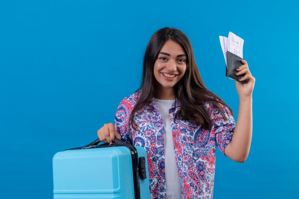 beautiful tourist woman holding travel suitcase and passport with tickets looking at camera with smile on face happy and positive travel concept standing over blue background
